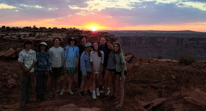 A group of students smile for a group photo while the sun sets behind them.